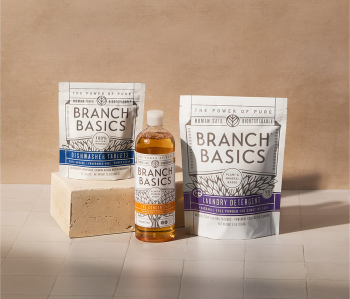 A bottle of Branch Basics Concentrate, a pouch of Dishwasher Tablets, and a pouch of Laundry Detergent are displayed on tiled surface and neutral background.
